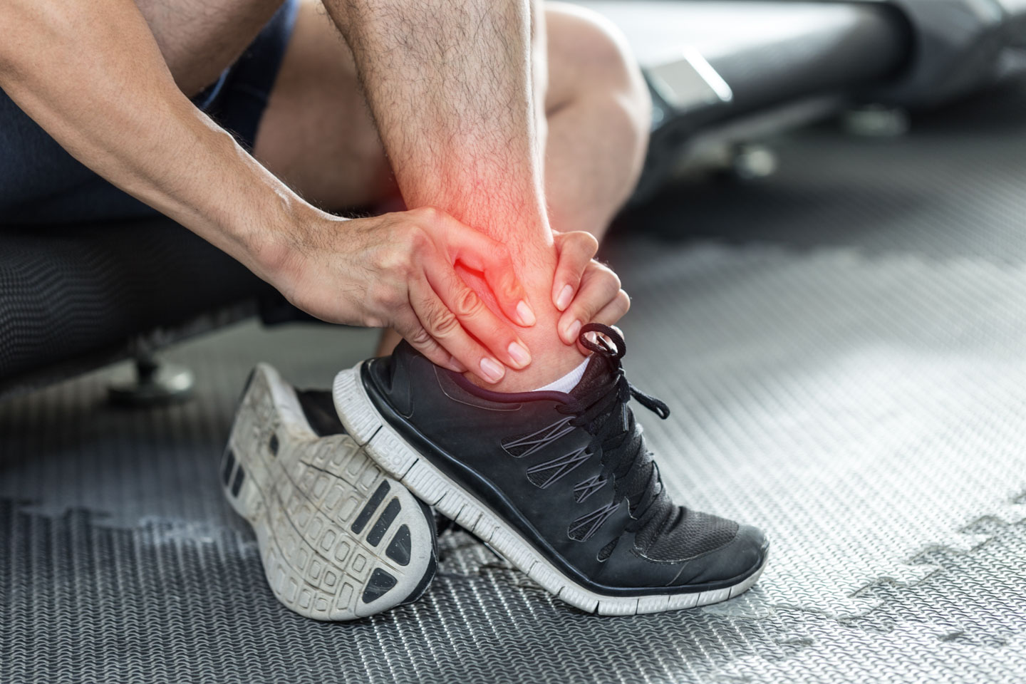 Person sitting on gym floor holding their injured ankle with a red highlight indicating pain.