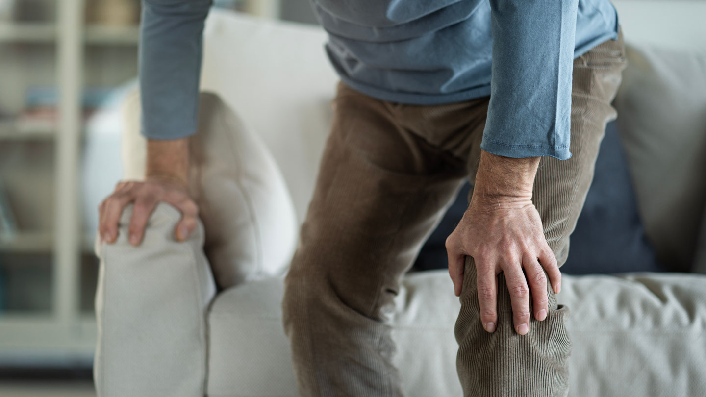 Person in brown pants holding knee and a sofa armrest, suggesting knee pain or discomfort.